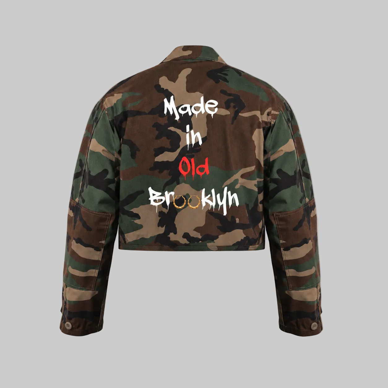 A cropped camouflage jacket shown from the back on a neutral gray background, featuring the text 'Made in Old Brooklyn' printed across the back in white, red, and gold lettering, with the o's in Brooklyn.replaced with hoop bamboo earrings.